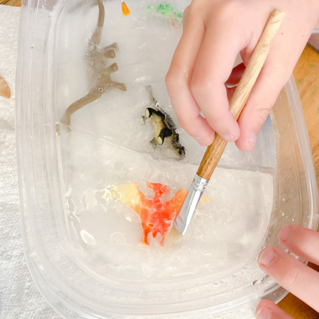 Toy dinosaurs frozen in ice. Hand with brush and water.